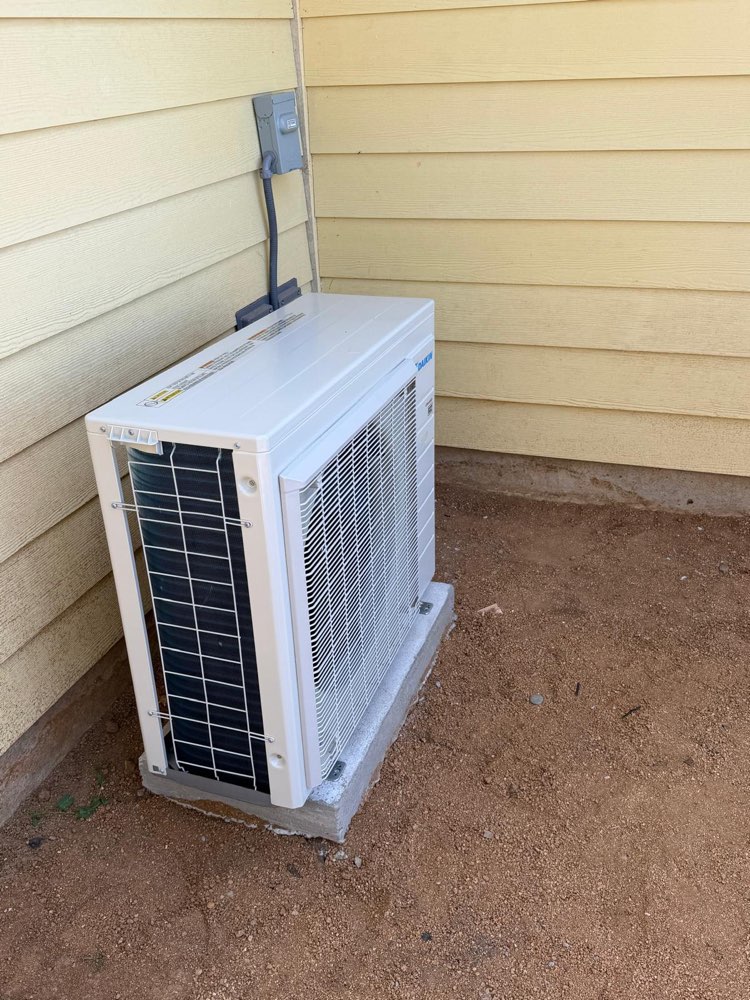 White Daikin outdoor mini-split unit on a concrete pad beside yellow siding.