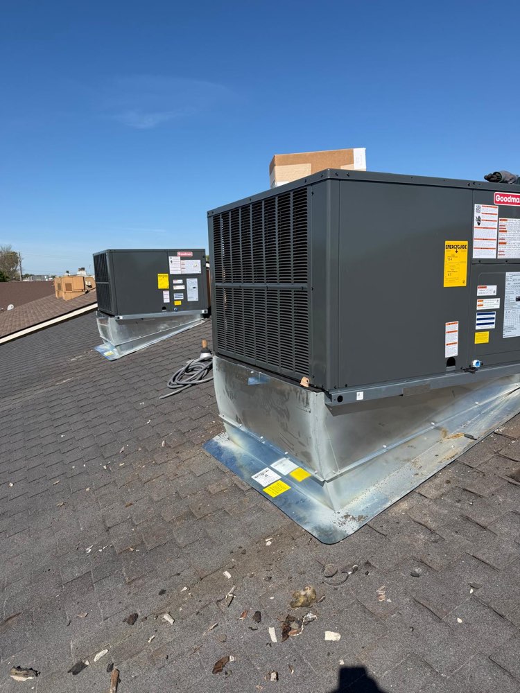 Two gray Goodman packaged HVAC units on raised metal bases on a shingle roof.