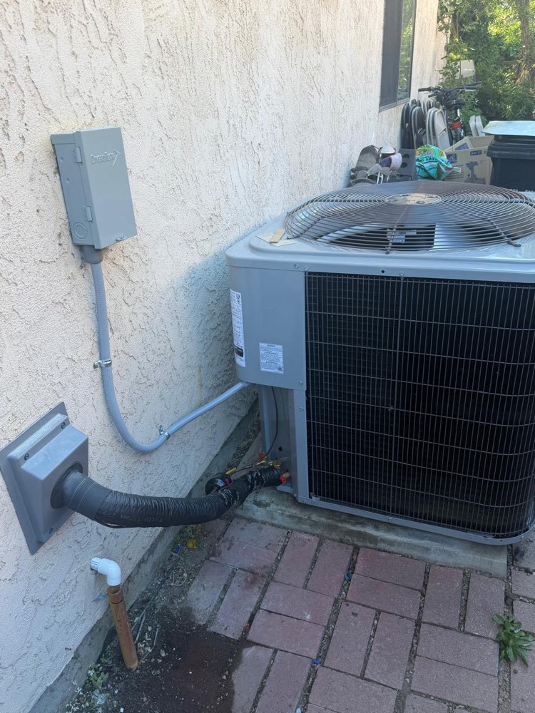 Outdoor air-conditioning unit beside a stucco wall, with insulated lines and an electrical connection visible.