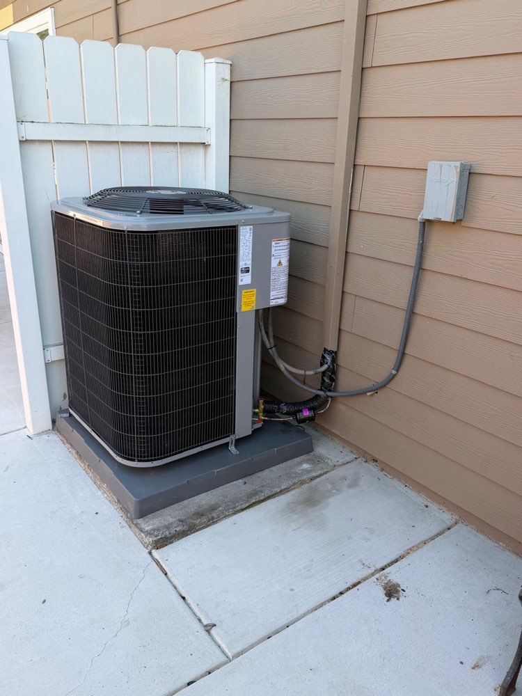 Outdoor air-conditioning unit on a base between a white fence and a siding wall.